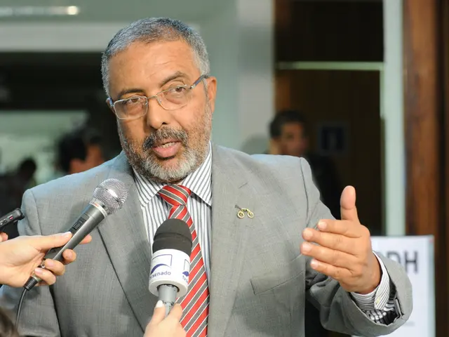 In this image there is a man, he is wearing suit and talking to journalists who are holding mics in...